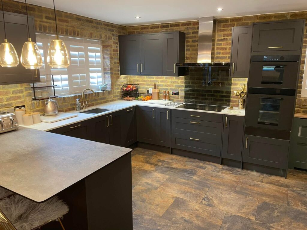 Modern kitchen with dark cupboards, exposed brick walls, under-cupboard lighting, built-in oven, and pendant lights over a breakfast bar. There are various small appliances and utensils on the worktops.
