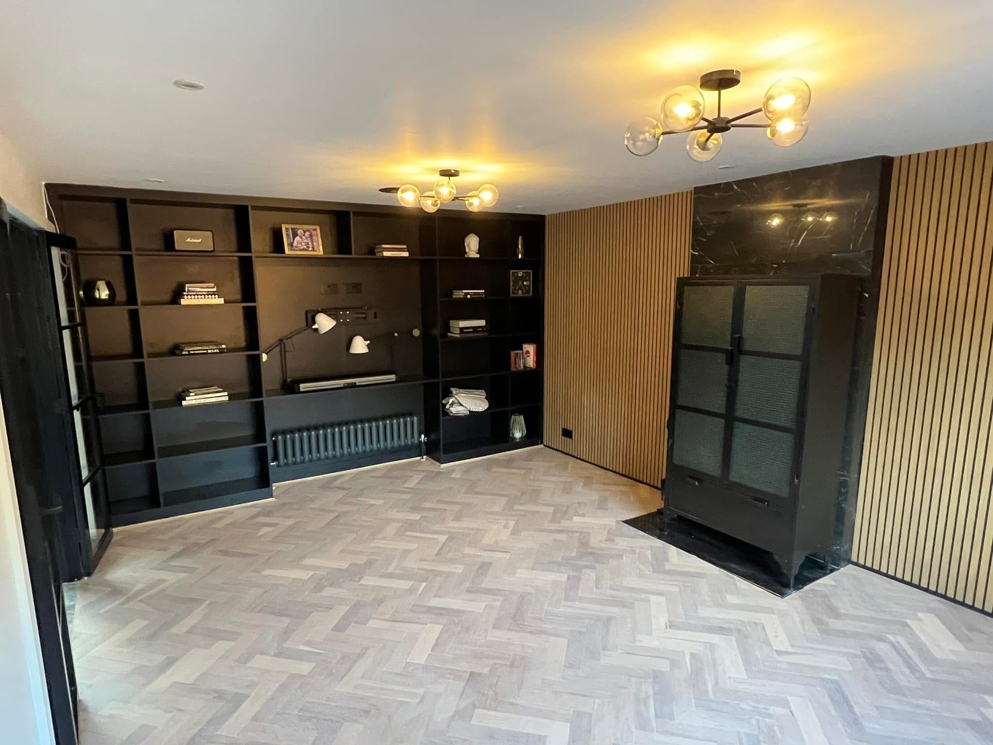 A modern living room with light herringbone wood flooring, black built-in shelves with books and decor, a black cabinet with glass doors, marble panelling, wood slat walls, and contemporary ceiling lights.