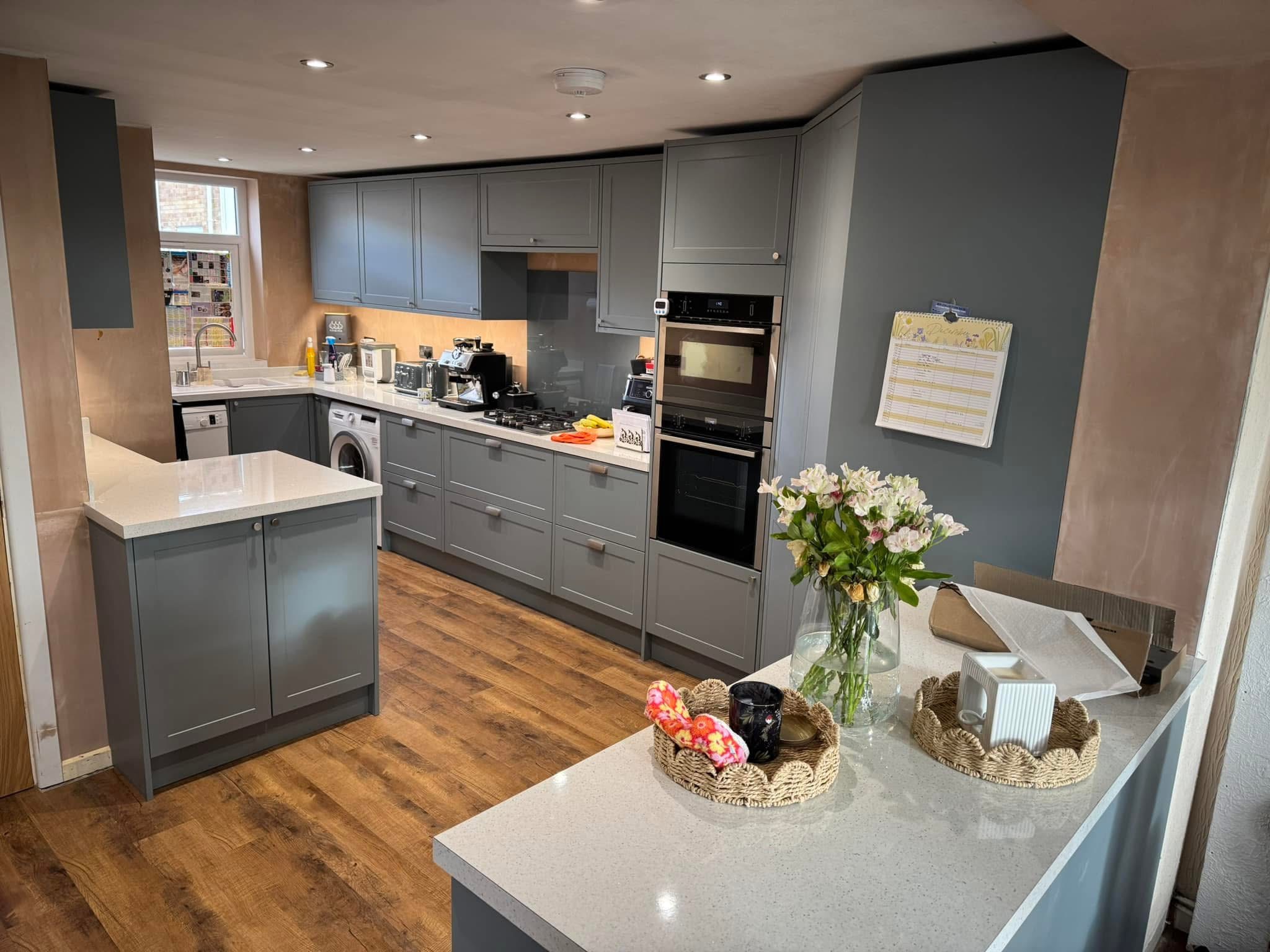 Modern kitchen with grey cupboards, wooden flooring, and white worktops. Fresh flowers and decorative trays are on the island, and appliances such as a washing machine, oven, and microwave are visible along the back wall.