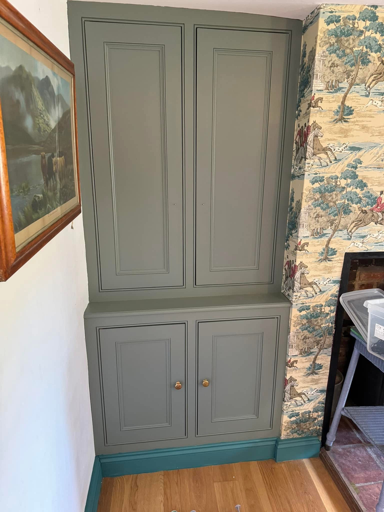 A sage green fitted cupboard with two upper and two lower doors, set in a corner next to wallpaper with a nature scene and a wooden floor. A framed landscape painting hangs on the adjoining wall.