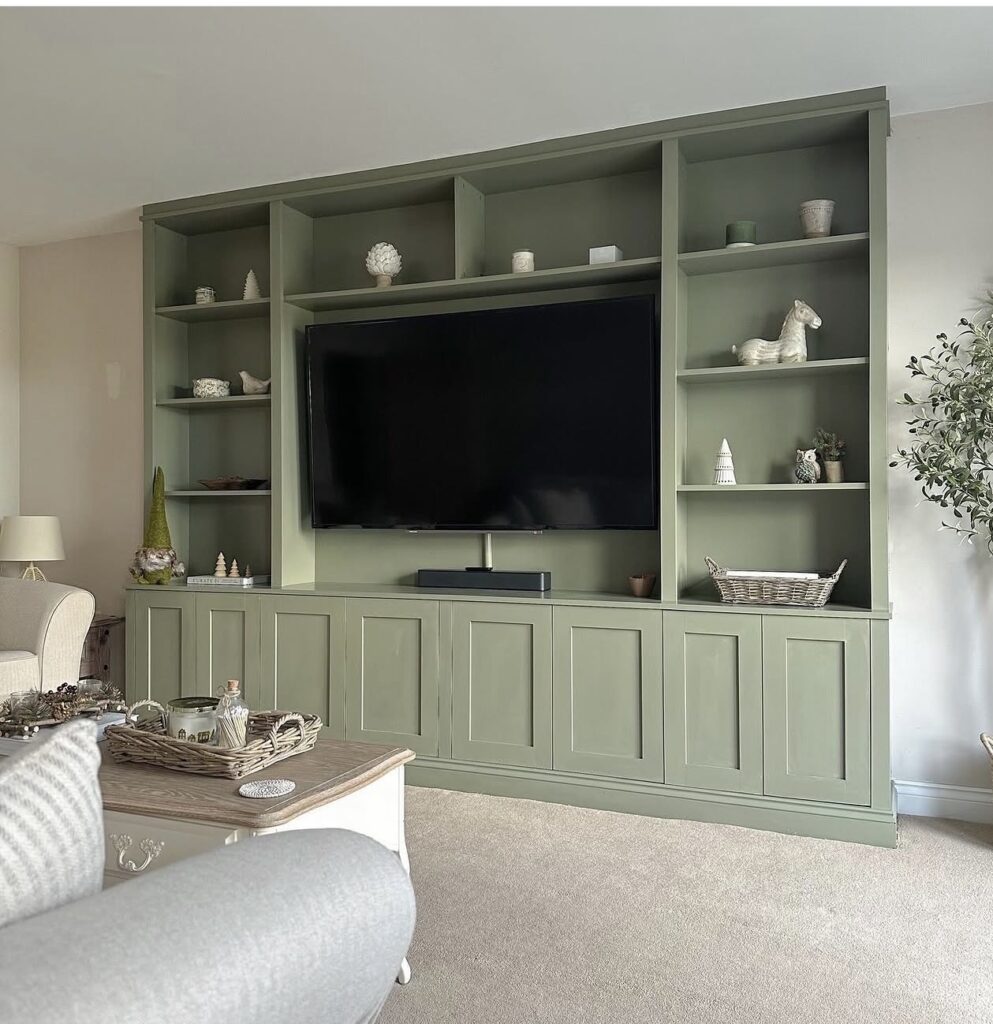Large green built-in shelving unit with a flat-screen telly in the centre, decorated with vases, figurines, baskets, and seashells, in a bright, tidy living room with neutral-coloured furniture and carpet.