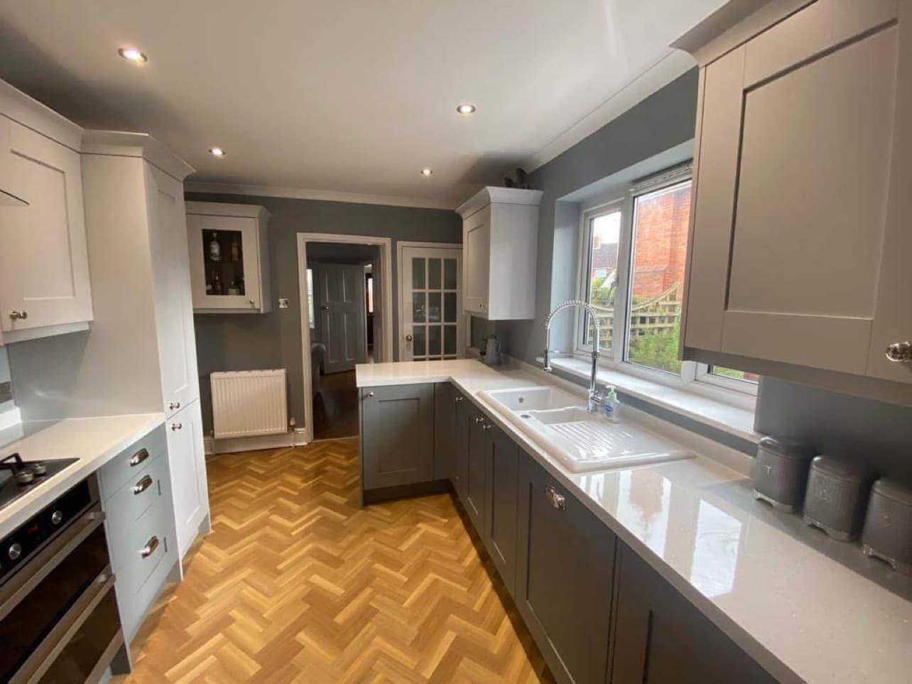 Modern kitchen with grey cabinets, white worktops, a built-in oven, herringbone wood floor, and a large window above the sink letting in natural light. The room is well-lit with ceiling spotlights.