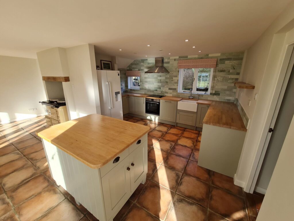 Bright modern kitchen with terracotta floor tiles, wooden worktops, an island in the centre, white cupboards, stainless steel appliances, and green tile splashback. Sunlight streams through windows onto the floor.