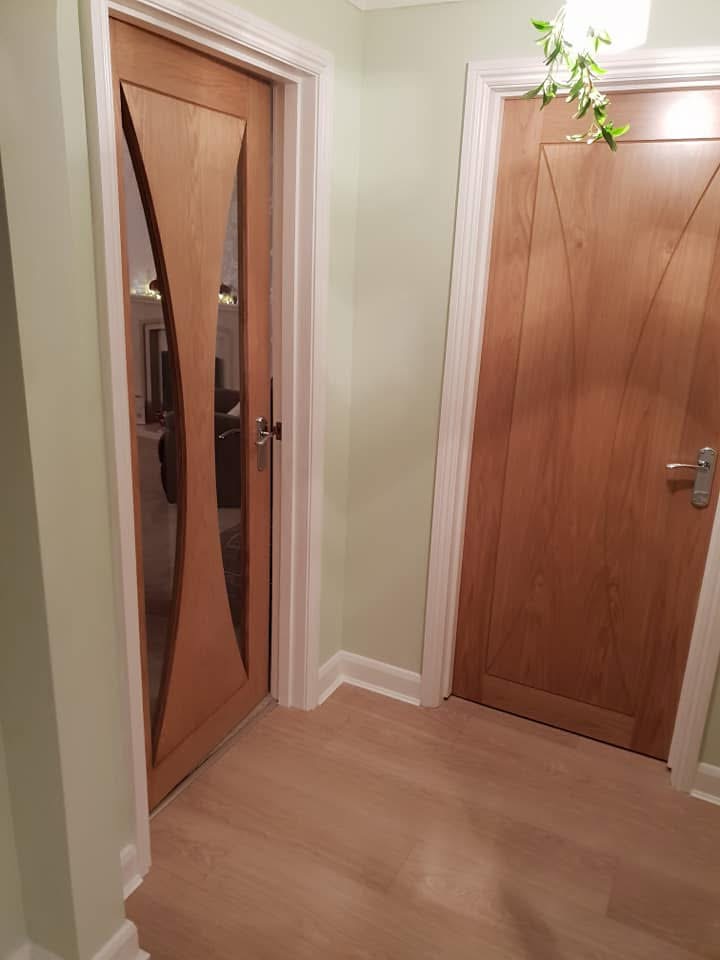 A corridor with light wood flooring, pale green walls, and two wooden doors; one door has a glass panel with curved wooden details, and the other is plain wood with a silver handle. A small plant hangs from the ceiling.