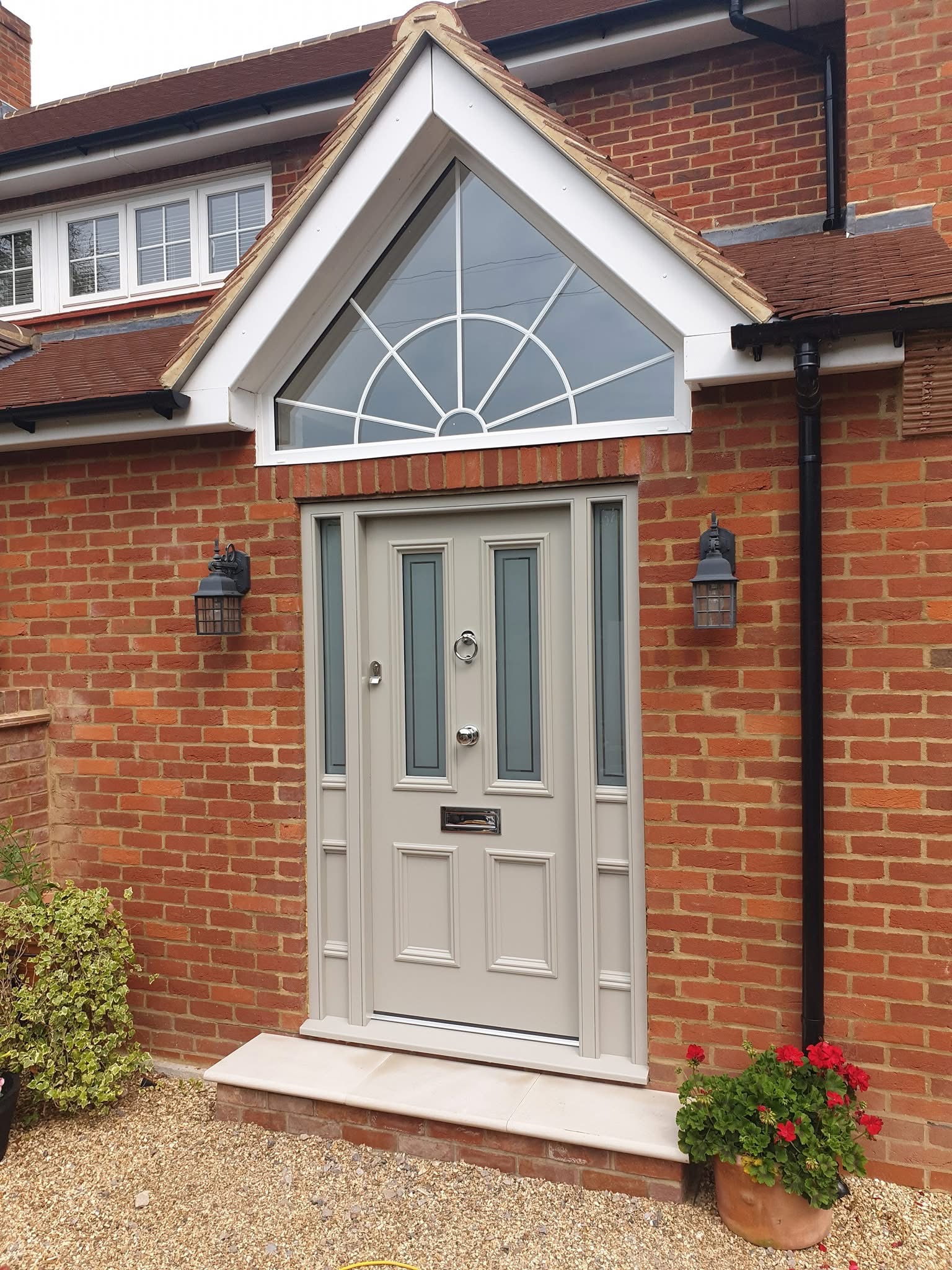 A light grey front door with glass panels and silver fittings, crafted by a skilled Basingstoke carpenter, graces a red brick house. Flanked by lanterns and topped with a triangular window, a potted plant with red flowers sits to the right.