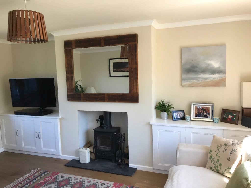 A cosy living room with a wood-burning stove, large rustic mirror above it, TV on a white cabinet, framed photos, a plant, and a beige sofa with patterned cushions against cream-coloured walls.