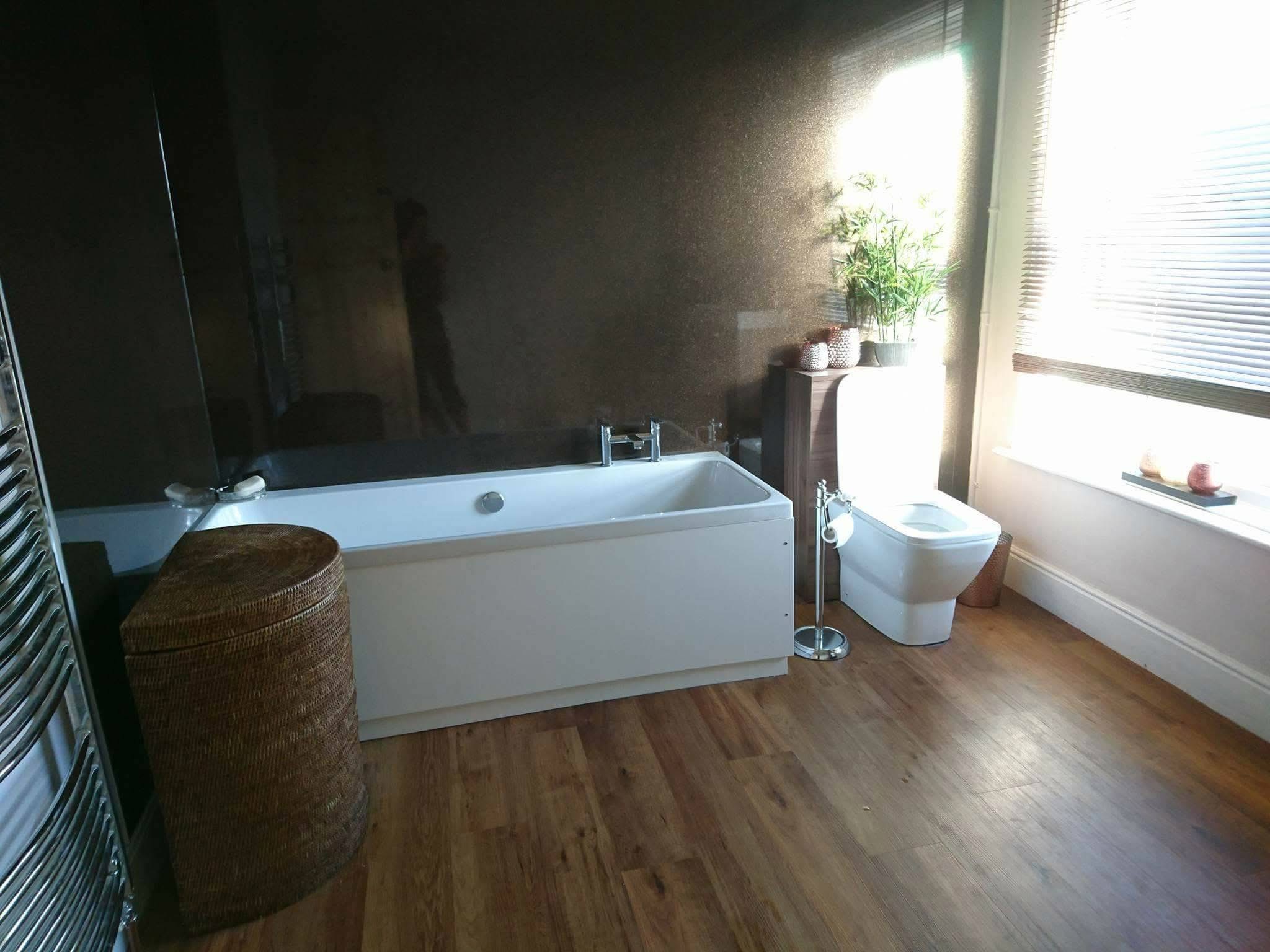 Modern bathroom featuring wood flooring crafted by a skilled Basingstoke carpenter, a white bath, wicker laundry basket, toilet, large window with blinds, and plants on a small shelf with sunlight streaming in.