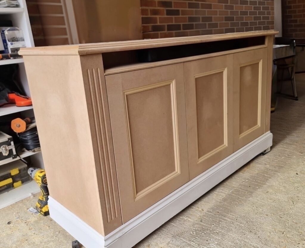 A partially finished wooden cupboard with decorative panels, crafted by a skilled Andover carpenter, stands on a white base in a workshop. The unpainted cupboard is surrounded by tools and shelves in the background.