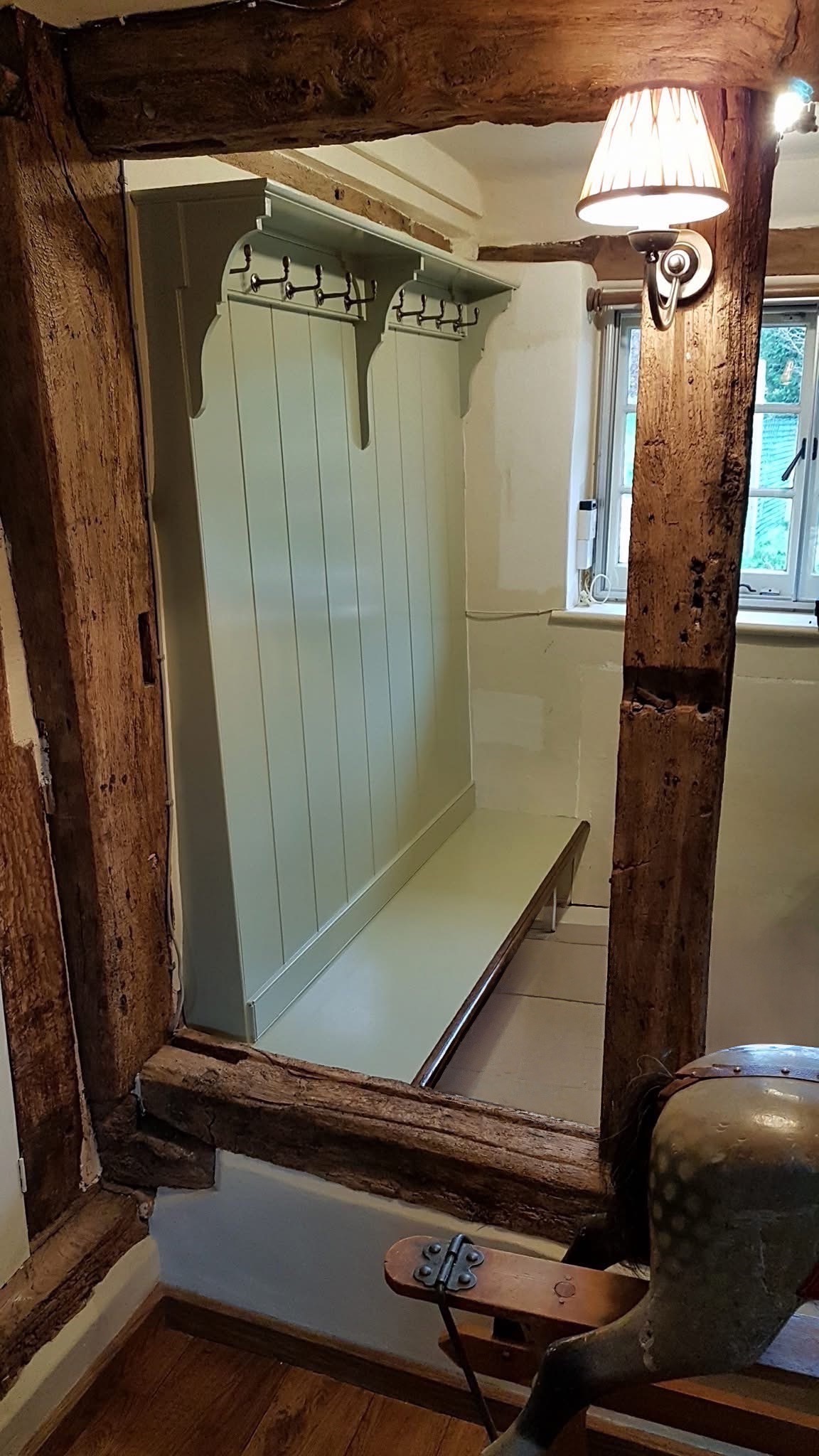 A rustic wooden-framed mirror, crafted by a skilled Andover carpenter, reflects a pale green bench with hooks above, set against a panelled wall. A lit wall sconce and lush greenery outside the window complete the charming scene.