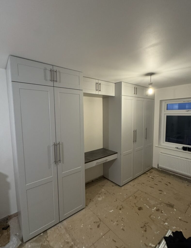 A room expertly fitted by a Basingstoke carpenter features new light grey wardrobes, a built-in desk, and upper cupboards. The unfinished floor is covered with protective sheets and paint splatters. A window and radiator appear on the right.