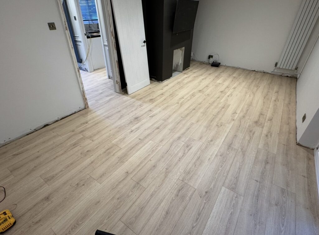A room with newly installed light wood laminate flooring, white walls, and minimal furniture. A Basingstoke carpenterโs tools are visible, hinting at ongoing renovation work through the doorway leading to another room.