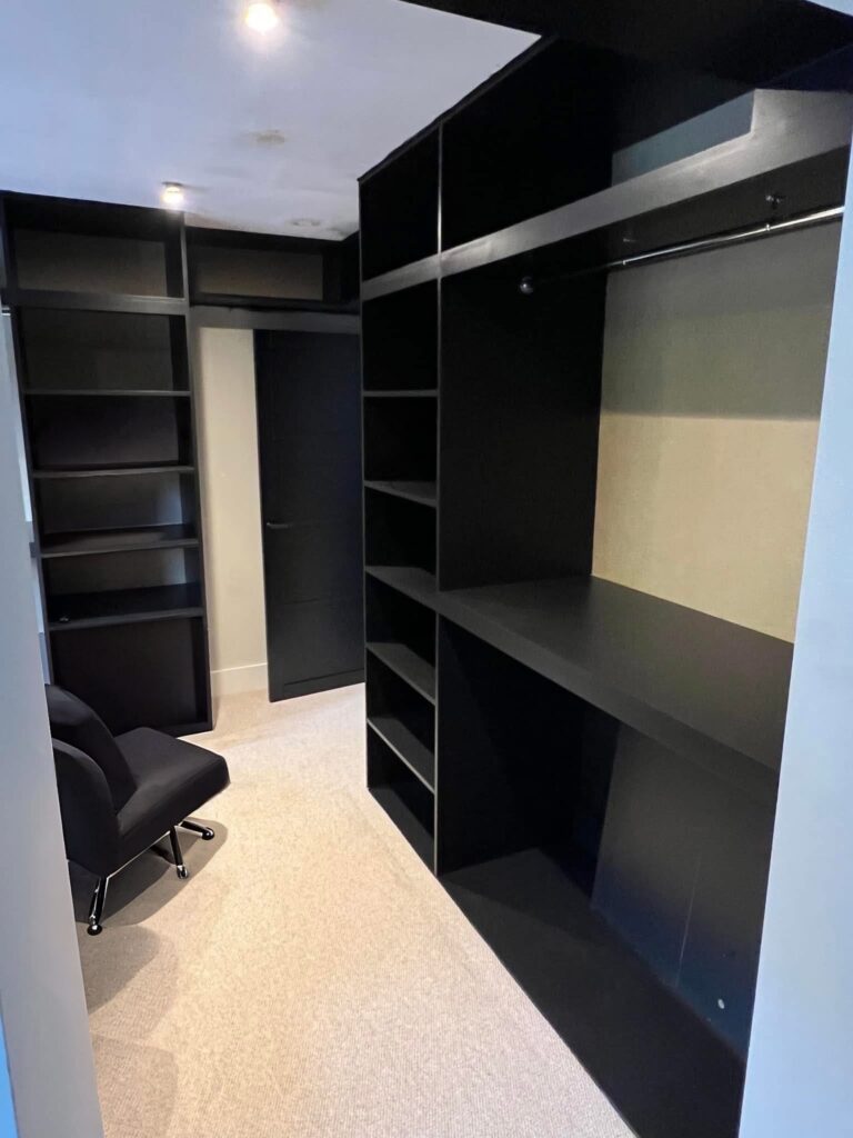 A modern walk-in wardrobe with black open shelves and hanging space, beige carpet flooring, a black chair, and overhead lighting—expertly crafted by a skilled Andover carpenter.