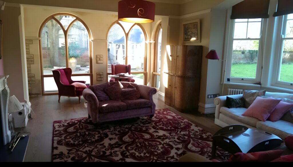 A cosy living room crafted by a skilled Andover carpenter features arched windows, a purple sofa, red armchairs, a white sofa, and a patterned rug. Sunlight streams in, highlighting the warm atmosphere and garden view.