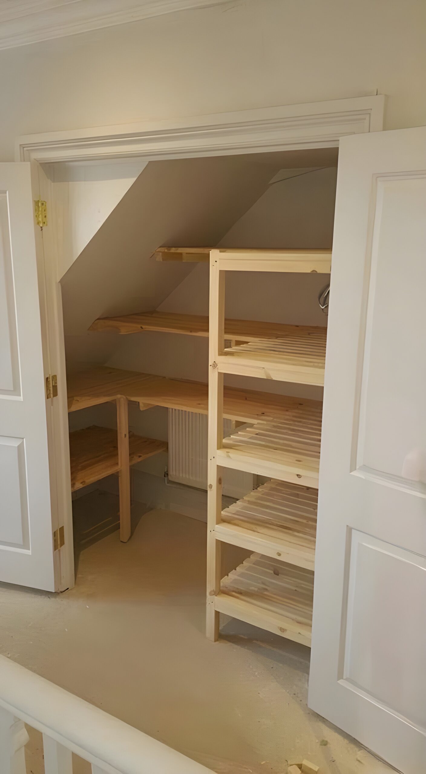 A cupboard with open double doors reveals wooden shelves and corner shelving units built under a sloped ceiling, maximising storage space in a small, well-lit room.